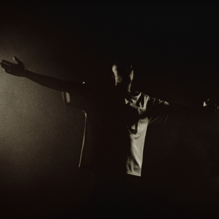 En person står med armene udstrakt i et mørkt, tåget rum med dramatisk belysning, der kun delvist oplyser ansigt og tøj. Den stærke kontrast mellem lys og skygge skaber en intens og teatralsk stemning.