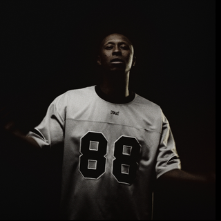 En mand står i et mørkt rum iført en lys Everlast sportstrøje med tallet "88" tydeligt trykt på fronten. Han er svagt oplyst forfra, hvilket skaber en dramatisk kontrast mellem lys og skygge, mens han kigger roligt frem med armene let ud til siden.