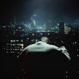 En mand set bagfra står foran et vindue med udsigt over en oplyst storby om natten. Han bærer en lyse trøje og har armene løftet, hvilket giver en dramatisk silhuet mod den glitrende skyline i mørket. Stemningen er filmisk og intens.