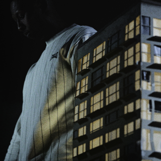 En mand i en lysegrå, ribbet Everlast-trøje står i et mørkt rum ved siden af en oplyst arkitekturmodel af en boligbygning. Lyset fra modellen kaster skygger og varme toner på hans trøje, hvilket skaber en dramatisk kontrast mellem menneske og bystruktur.