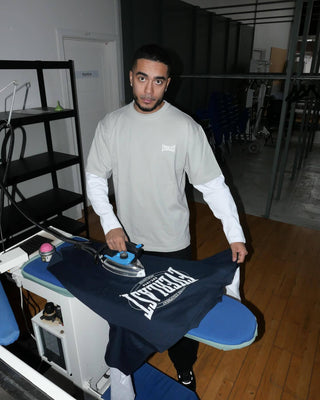 En mand står ved et strygebord og presser et stort hvidt Everlast-logo på en mørk t-shirt med et strygejern. Han er iført en lysegrå Everlast t-shirt over en hvid langærmet trøje og kigger direkte mod kameraet i et industrielt rum med trægulv.