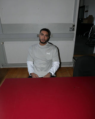 En mand sidder ved et bord med rød overflade iført en lysegrå Everlast t-shirt med hvidt logo over en hvid langærmet trøje. Han kigger roligt mod kameraet i et industrielt rum med trægulv og en radiator bag sig.