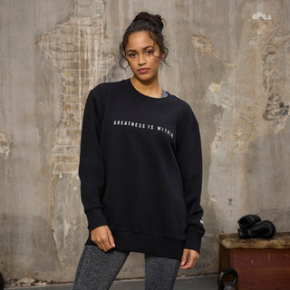 En kvinde står foran en slidt betonvæg iført en sort sweatshirt med teksten "GREATNESS IS WITHIN" trykt på brystet. Hun har håret opsat og kombinerer trøjen med mørke træningsleggings, hvilket giver et afslappet og sporty look.