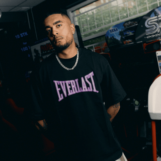 En mand står i et arcadecenter iført en sort Everlast t-shirt med stort lilla logo på brystet og en sølvkæde om halsen. Han poserer med armene bag ryggen og et roligt, selvsikkert udtryk foran farverige spillemaskiner.