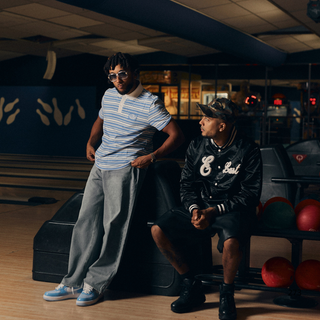 To mænd befinder sig i en bowlinghal omgivet af kugler og baner. Den ene står iført en lyseblå og hvidstribet polo, posede grå bukser og blå sneakers, mens den anden sidder iført en sort baseballjakke med "E" og "Last" skrevet på samt shorts og sneakers. Stemningen er afslappet med et sporty streetwear-look.