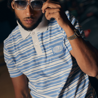 En mand poserer i en bowlinghal iført en blå- og hvidstribet Everlast polo med hvid krave. Han justerer sine transparente solbriller med den ene hånd og bærer et armbåndsur, hvilket giver et selvsikkert og stilbevidst udtryk.