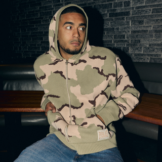 En mand sidder i en mørk loungestemning iført en lysegrøn og beige camouflagemønstret Everlast hættetrøje med hætten oppe. Han har blå jeans på og kigger til siden med et eftertænksomt udtryk, mens han hviler armene afslappet på bænken.