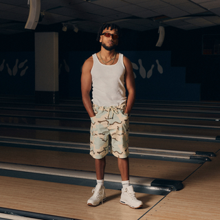 En mand står midt på en bowlingbane iført en hvid tanktop, lyse camouflagemønstrede shorts og hvide sneakers. Han har solbriller på og hænderne i lommerne, hvilket giver et afslappet og selvsikkert sommerlook i en indendørs, sporty setting.