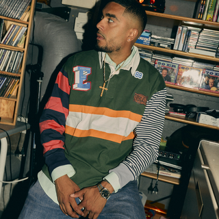 En mand sidder i et rum fyldt med cd’er og lydudstyr iført en farverig rugby-inspireret trøje med striber i grøn, orange, hvid og navy samt forskellige logoer og lapper, herunder et Everlast-mærke. Han bærer accessories som guldkæde og ur og kigger til siden med afslappet attitude.