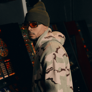 En mand står i dæmpet belysning foran blinkende spillemaskiner iført en beige og grøn camouflagemønstret hættetrøje. Han har orange tonede solbriller og en olivengrøn hue på, hvilket giver et cool og urban look i arcade-miljøet.