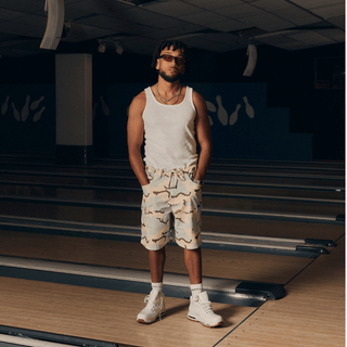 En mand står midt på en bowlingbane iført en hvid tanktop, lyse camouflagemønstrede shorts og hvide sneakers. Han bærer solbriller og har hænderne i lommerne, hvilket giver et afslappet og selvsikkert udtryk i den dæmpede hal.