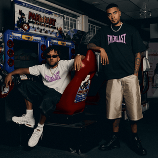 To mænd poserer foran en retro Mario Kart arkademaskine, begge iført oversized Everlast t-shirts med lilla logo. Den ene sidder i en rød racersæde iført hvid t-shirt, solbriller og sneakers, mens den anden står ved siden af i sort t-shirt og beige shorts, hvilket skaber et cool og nostalgisk streetwear-look.