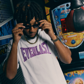 En mand justerer sine solbriller foran en boksemaskine i et arkadecenter iført en hvid Everlast t-shirt med stort lilla logo. Han bærer også en guldkæde og ur, hvilket understreger det afslappede og stilbevidste streetwear-look.