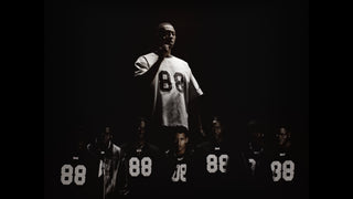 En dramatisk scene viser en mand oplyst fra oven iført en lys Everlast trøje med tallet "88", omgivet af flere personer i mørke med samme trøje. Den stærke kontrast mellem lys og skygge skaber en intens, næsten teatralsk stemning med fokus på individualitet og fællesskab.