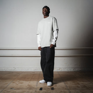 En mand står afslappet i et råt, industrielt rum iført en lysegrå Everlast t-shirt over en hvid langærmet trøje. Han bærer posede sorte bukser og hvide sneakers, og hans posering understreger et moderne streetwear-look.