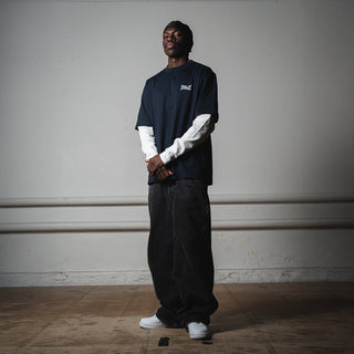 En mand står i et minimalistisk rum med hænderne samlet iført en mørkeblå Everlast t-shirt med hvidt logo over en hvid langærmet trøje. Han bærer posede sorte bukser og hvide sneakers, og poseringen fremhæver det afslappede streetwear-look.