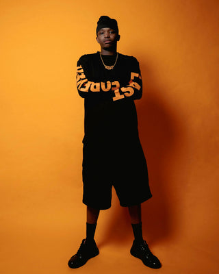 En mand står med armene over kors foran en orange baggrund iført sort streetwear med store orange "EVERLAST" tryk på ærmerne. Han har en sort hue, guldkæde, lange shorts og sorte sneakers, hvilket giver et selvsikkert og afslappet look.