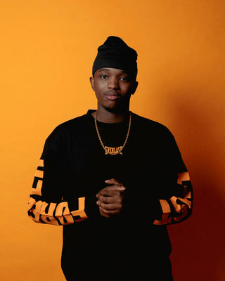 En mand står foran en orange baggrund iført en sort langærmet Everlast-trøje med store orange logoer på begge ærmer. Han har en sort hue på og bærer en guldkæde med et Everlast-navneskilt, mens han kigger roligt mod kameraet med hænderne foldet foran sig.