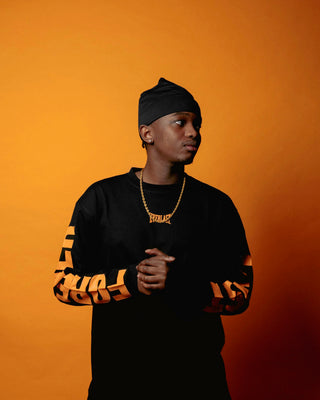 En mand står foran en orange baggrund iført en sort Everlast langærmet trøje med orange tryk på ærmerne, hvor der står "FOREVER". Han bærer en sort hue og en guldkæde med Everlast-logo, og han kigger til siden med hænderne samlet foran sig.