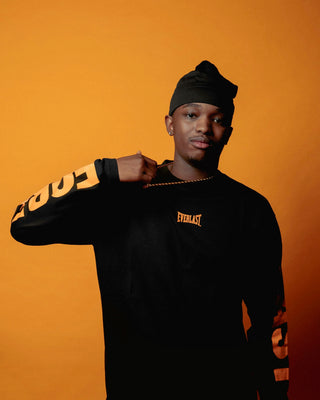 En mand poserer foran en orange baggrund iført en sort Everlast-trøje med orange logo på brystet og teksten "FOREVER" trykt ned ad ærmerne. Han holder en guldkæde med Everlast-navnet op mod brystet og bærer en sort hue, mens han kigger selvsikkert mod kameraet.