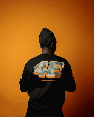 En mand står med ryggen til foran en orange baggrund iført en sort langærmet trøje med stort metallisk "4E" tryk på ryggen og teksten "BROOKLYN, NEW YORK" nedenunder. Han har en sort hue på og en guldkæde, hvilket fremhæver det urbane streetwear-look.