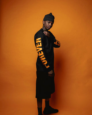 En mand poserer i profil foran en orange baggrund iført en sort langærmet trøje med ordet "FOREVER" trykt i store orange bogstaver ned ad ærmet. Han bærer en sort hue, sorte shorts og sorte sko, og hans attitude udstråler selvtillid og streetwear-stil.