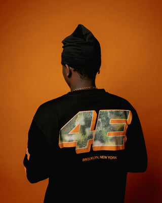 En mand står med ryggen til foran en orange baggrund iført en sort langærmet trøje med stort camouflagefarvet “4E” tryk på ryggen og teksten “BROOKLYN, NEW YORK” i orange nedenunder. Han bærer en sort hue og guldkæde, hvilket understreger det urbane streetwear-look.